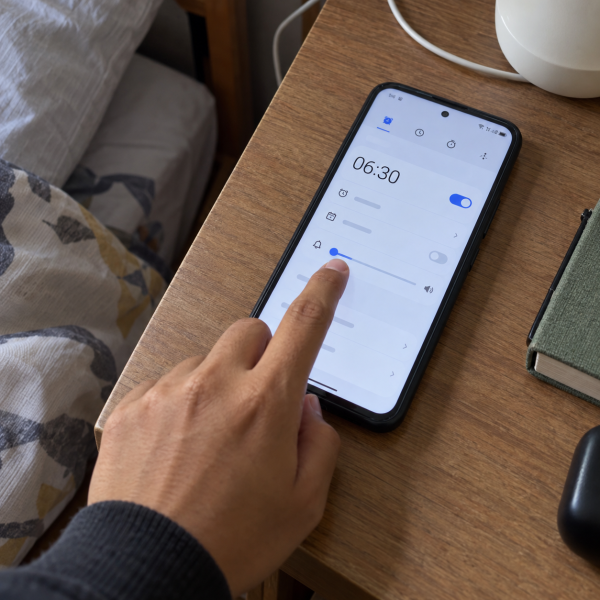 Tangan mengatur volume alarm pada smartphone yang menampilkan pengaturan alarm di meja samping tempat tidur.